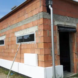 Budynek w trakcie prac elewacyjnych: widoczne ocieplenie fundamentów styropianem, ściany z czerwonej cegły z betonowymi wzmocnieniami, okna z szarymi ramami i metalowa rynna. Budynek jest w stanie...