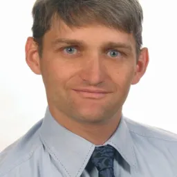 Portret mężczyzny w błękitnej koszuli i wzorzystym krawacie, uśmiechającego się do kamery na jasnym tle. Widoczne niebieskie oczy i starannie ułożone włosy.