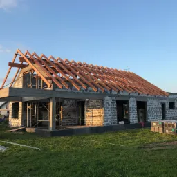 Surowy stan domu z pustak&oacute;w z drewnianą konstrukcją dachu w trakcie budowy, widoczne rusztowania i materiały budowlane na trawniku.