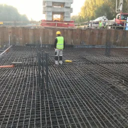 Pracownik w żółtym kasku i kamizelce odblaskowej stoi na zbrojeniu fundamentu, w tle szalunek, dźwig i betoniarka.