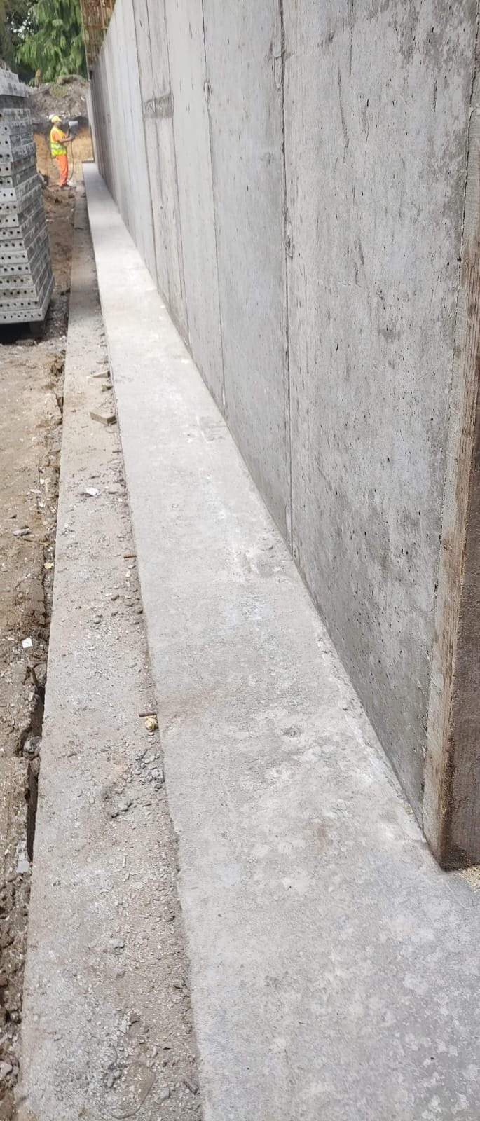 Szara ściana betonowa z widocznymi śladami deskowania, świeżo wylana, z pracownikiem w oddali, w tle zielona roślinność.
