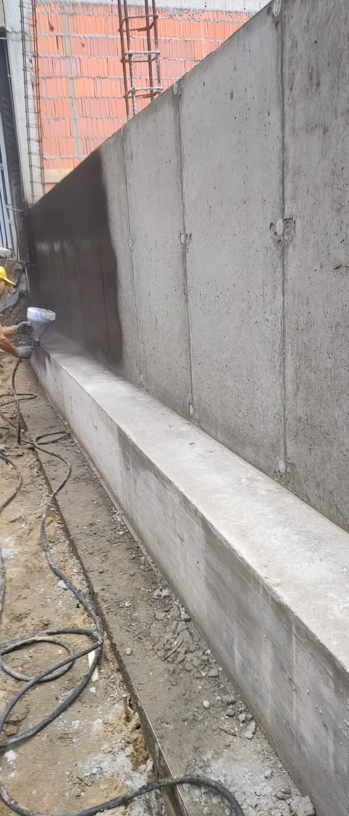 Natrysk hydroizolacji na betonową ścianę fundamentową, widoczny fragment ściany z cegieł oraz przewody elektryczne na ziemi.
