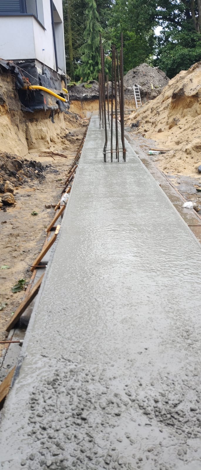 Świeżo wylany, długi fundament betonowy z wystającymi prętami zbrojeniowymi, otoczony wykopem i ziemią. Widoczny fragment budynku w tle.