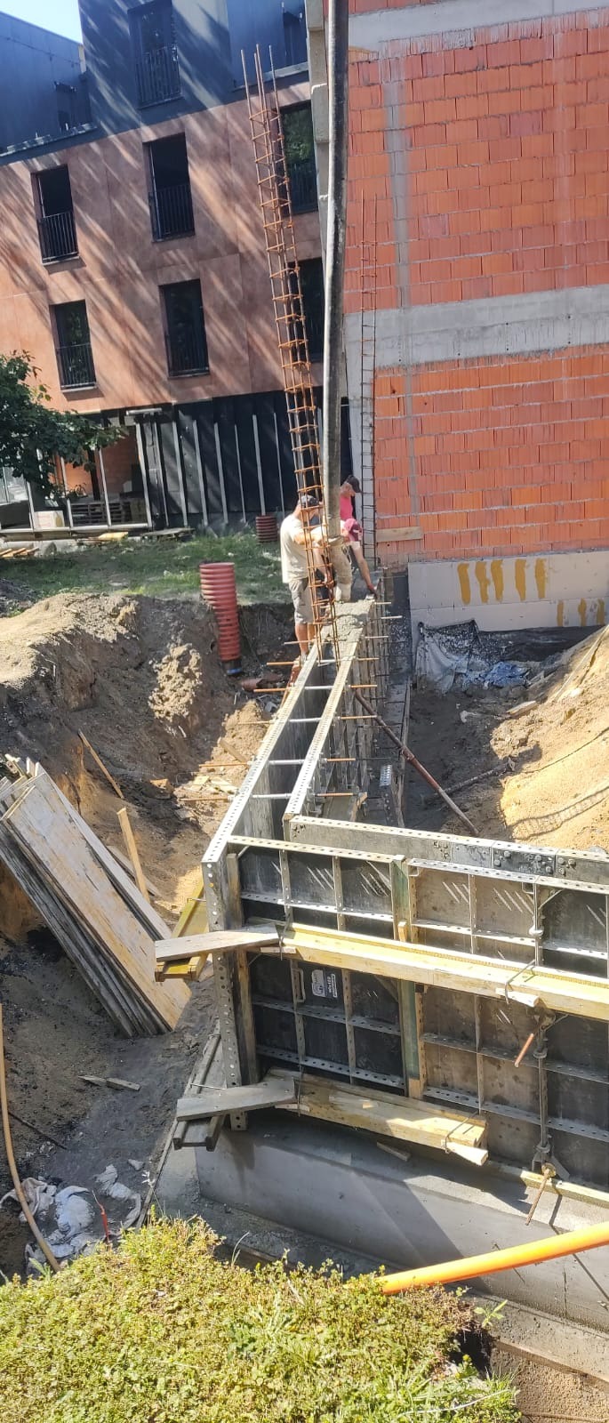 Wylewanie betonu do szalunków fundamentowych, widoczny betonowy wąż, pracownicy oraz zbrojenie. W tle budynek w trakcie wznoszenia.