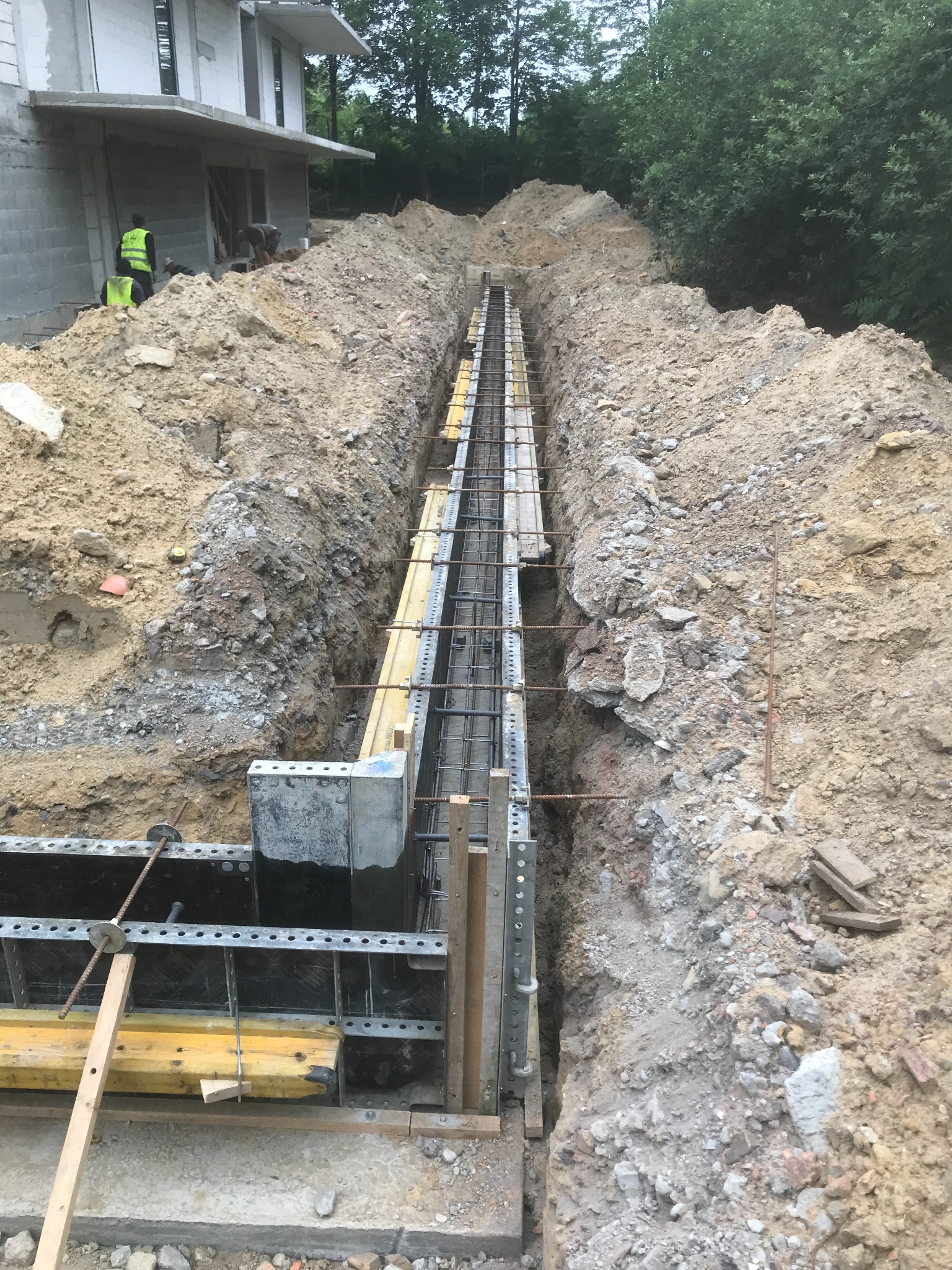 Wykop pod fundament z deskowaniem i zbrojeniem, widoczni pracownicy w odblaskowych kamizelkach, budynek w stanie surowym w tle.