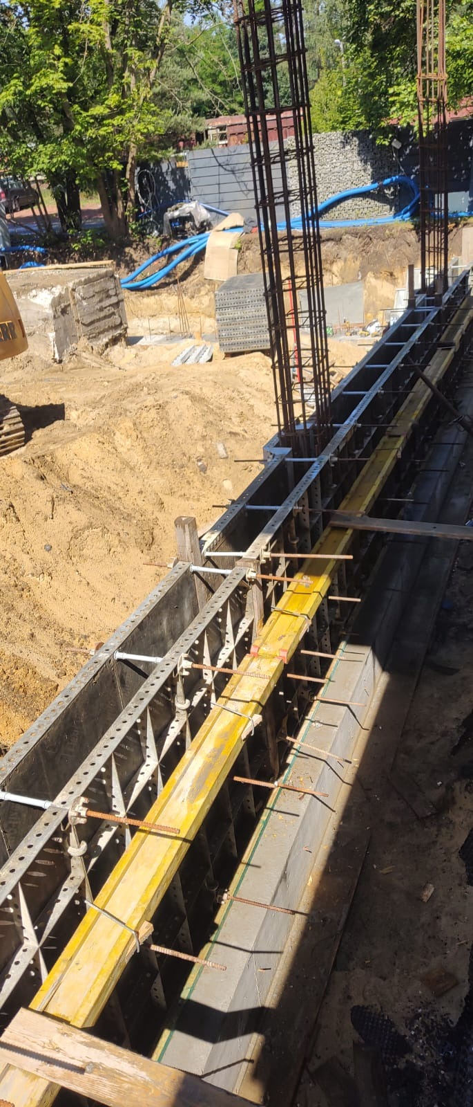 Szczegółowy widok szalunku fundamentowego z żółtymi deskami, zbrojenia pionowego i wykopu ziemnego na placu budowy w słoneczny dzień.