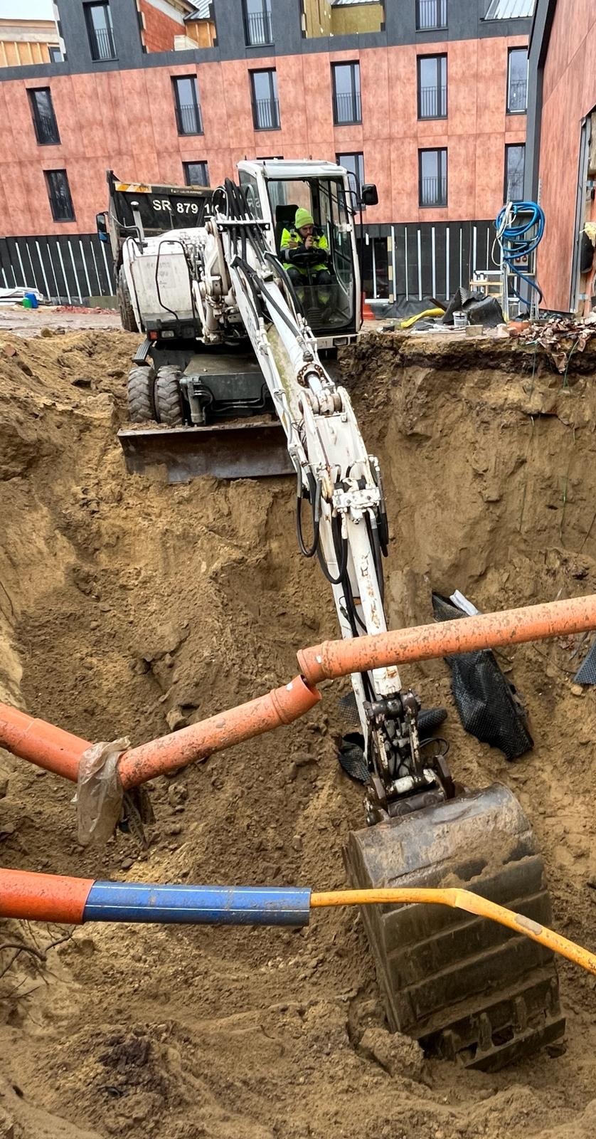 Biała koparka w głębokim wykopie z widocznymi pomarańczowymi rurami i niebieską osłoną kabla, operator w kabinie, budynek w tle.
