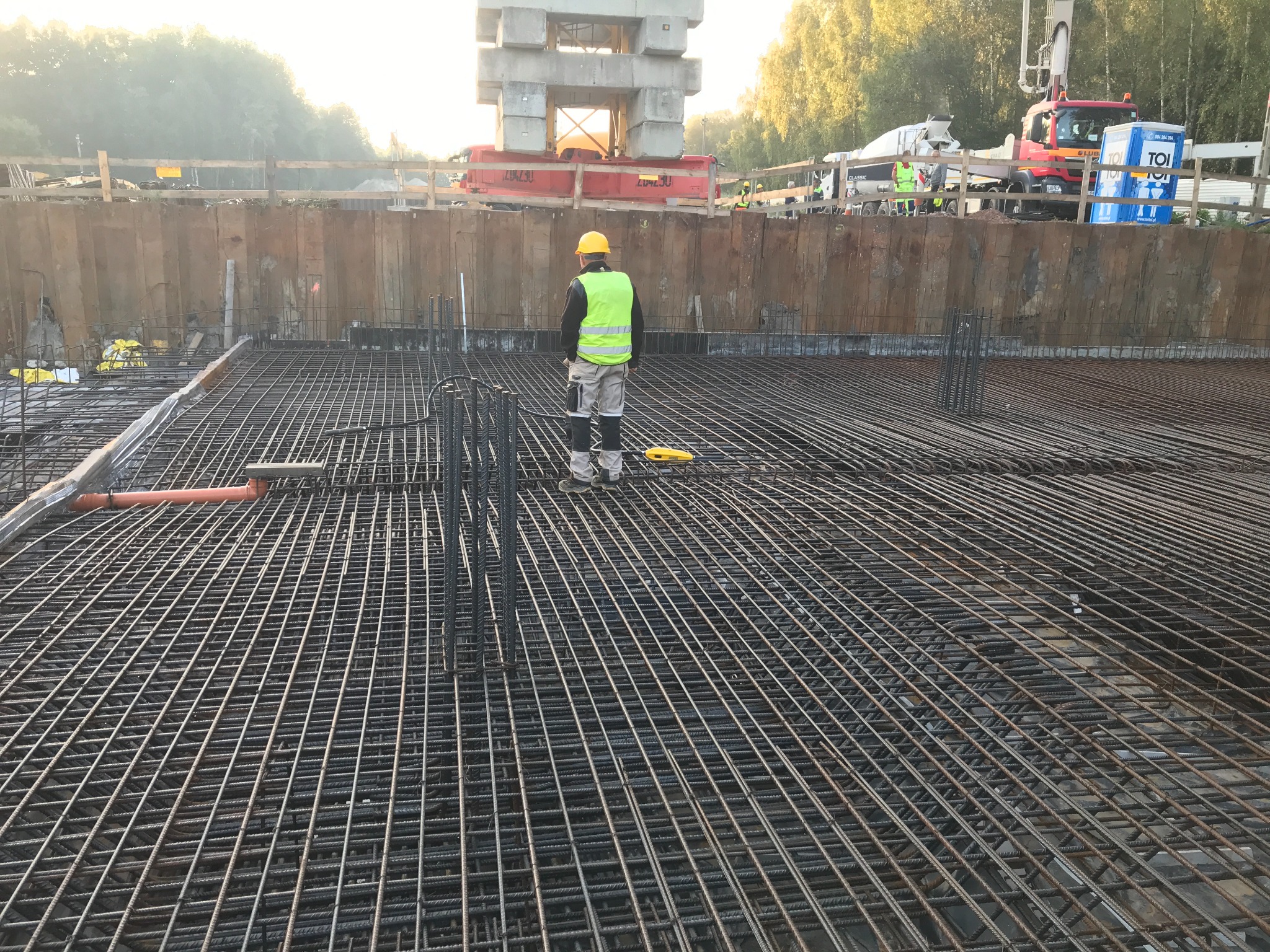 Pracownik w żółtym kasku i kamizelce odblaskowej stoi na zbrojeniu fundamentu, w tle szalunek, dźwig i betoniarka.