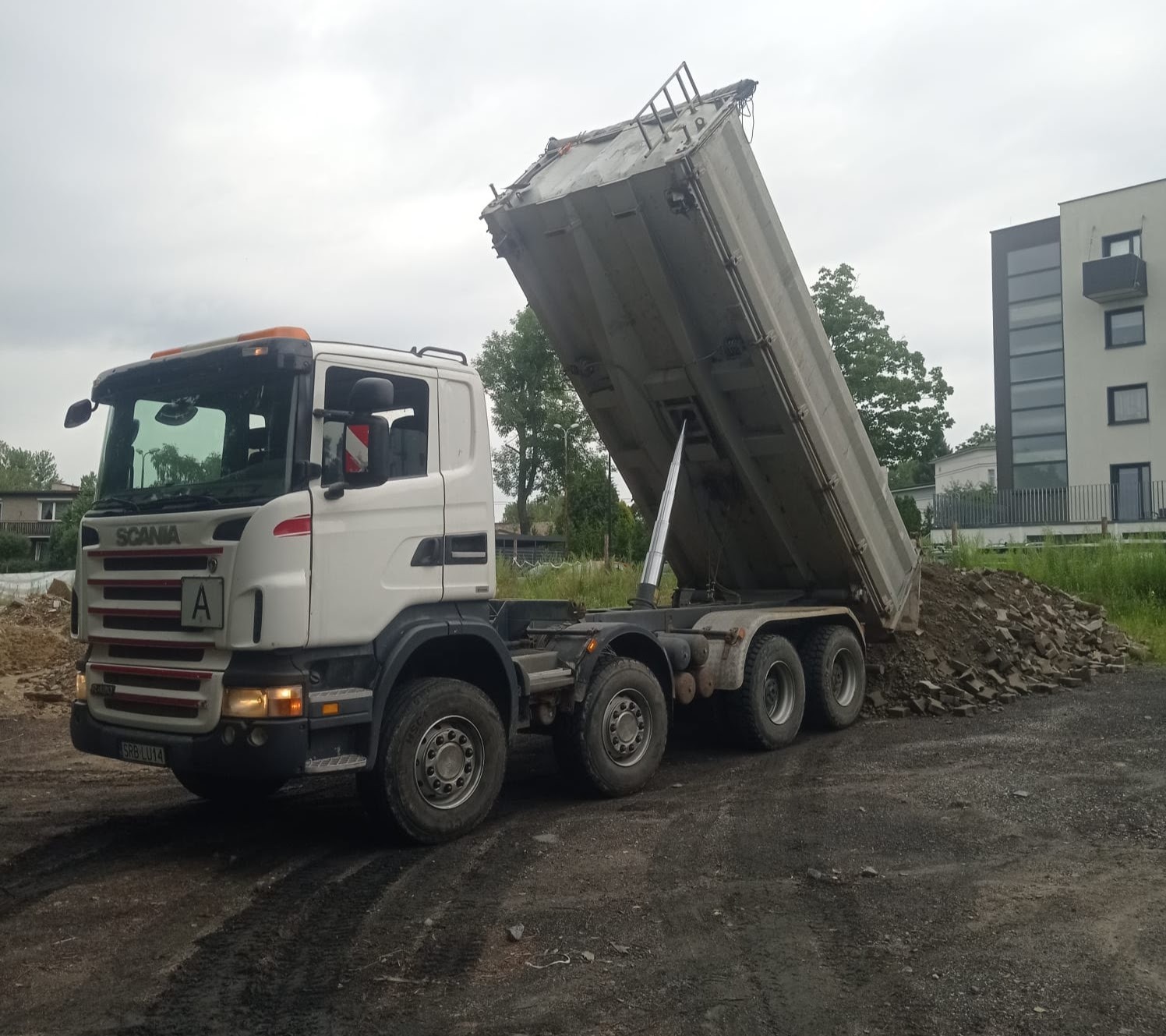 Wyładowywanie gruzu z wywrotki Scania na placu budowy, widoczny fragment budynku mieszkalnego w tle.