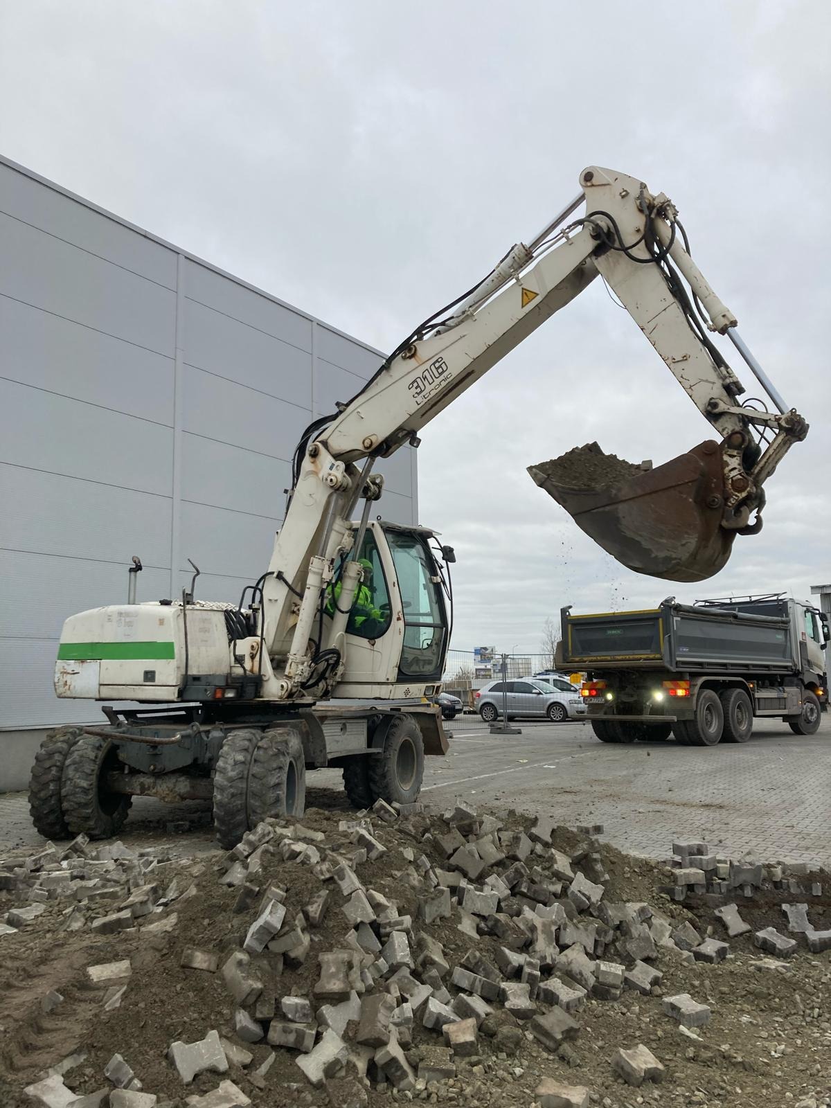 Biała koparka kołowa ładuje ziemię i gruz do wywrotki na tle szarego budynku magazynowego i pochmurnego nieba. Na pierwszym planie sterta potłuczonej kostki brukowej.