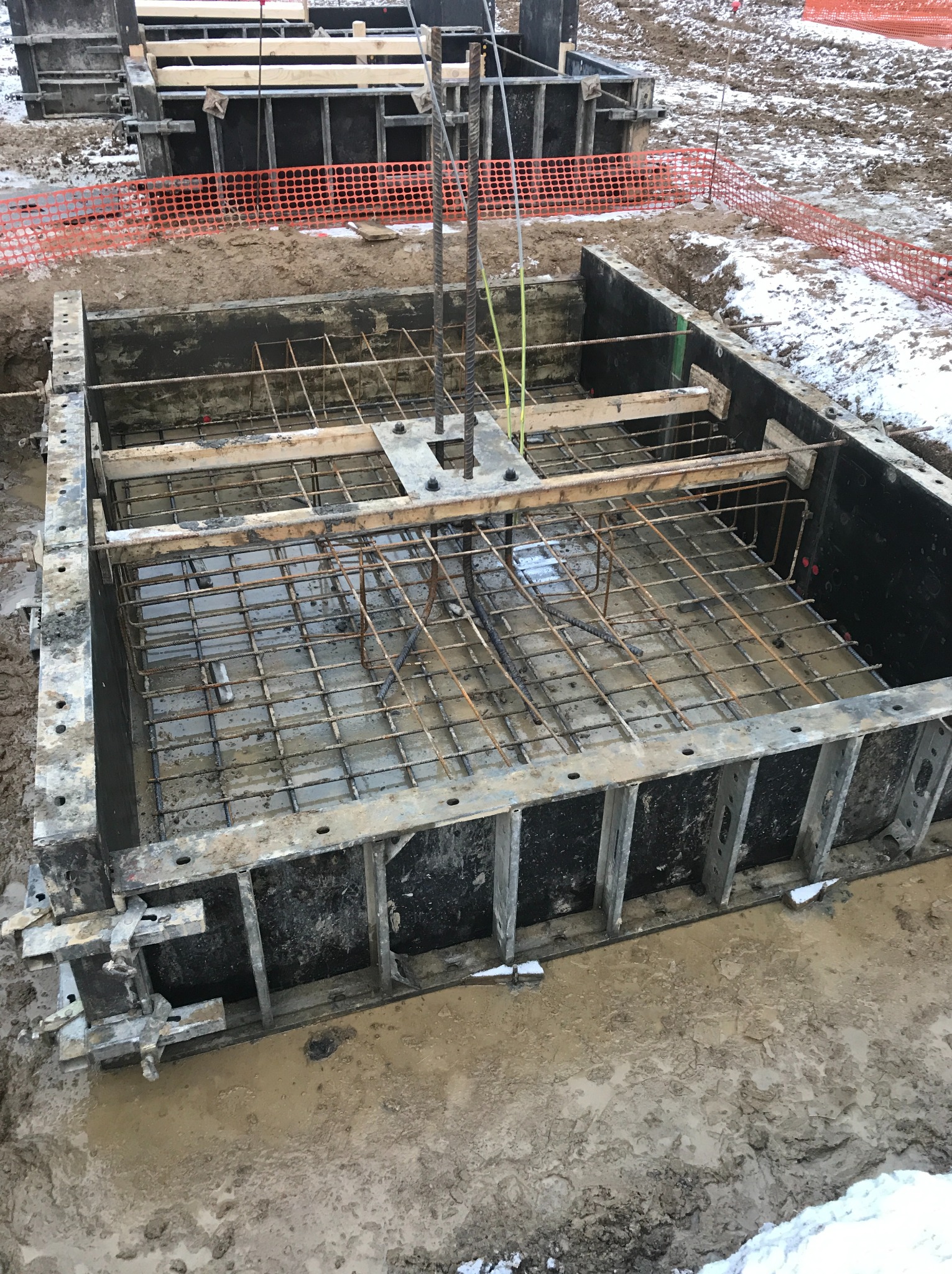 Szare szalunki fundamentowe zbrojone stalową siatką i drewnianymi belkami, częściowo wypełnione betonem, na placu budowy z resztkami śniegu.