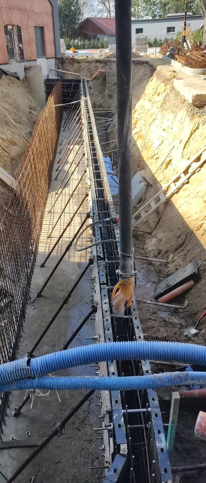 Wykop z deskowaniem i zbrojeniem przygotowany do wylewania betonu, widoczna pompa do betonu z żółtym wężem, niebieskie rury w wykopie.