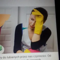 Zamyślona kobieta w fartuchu i żółtych rękawicach opiera głowę na dłoni, z widocznymi środkami czystości w tle, sugerując zmęczenie po sprzątaniu.