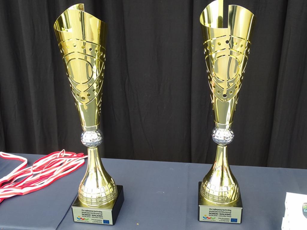 Dwie złote, błyszczące statuetki z ażurowymi elementami na czarnym tle, wręczone za konkurs Nordic Walking.