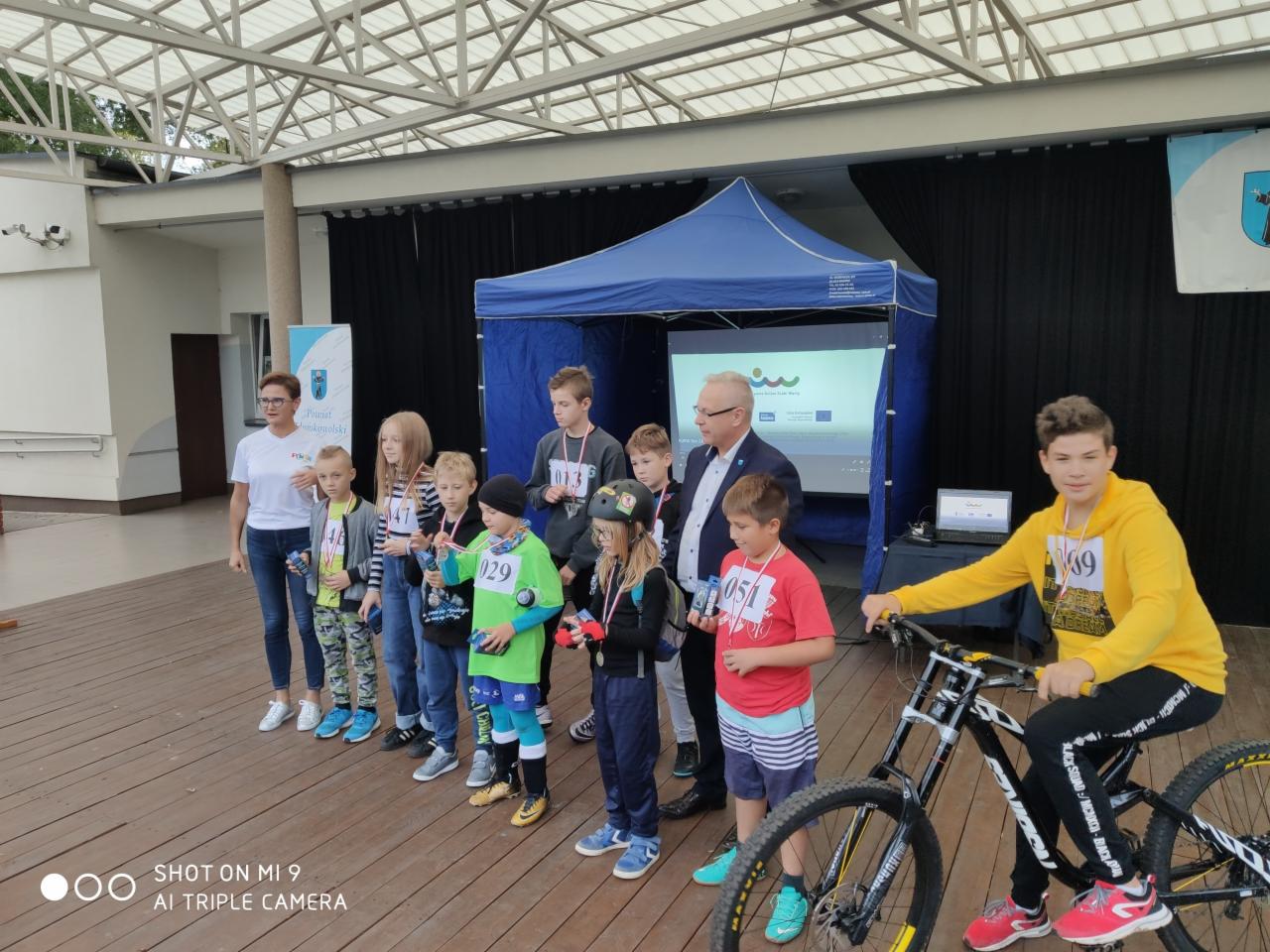 Grupa dzieci z medalami na szyi, stojących na scenie podczas ceremonii wręczenia nagród, z chłopcem na rowerze po prawej stronie i urzędnikiem w garniturze w tle.