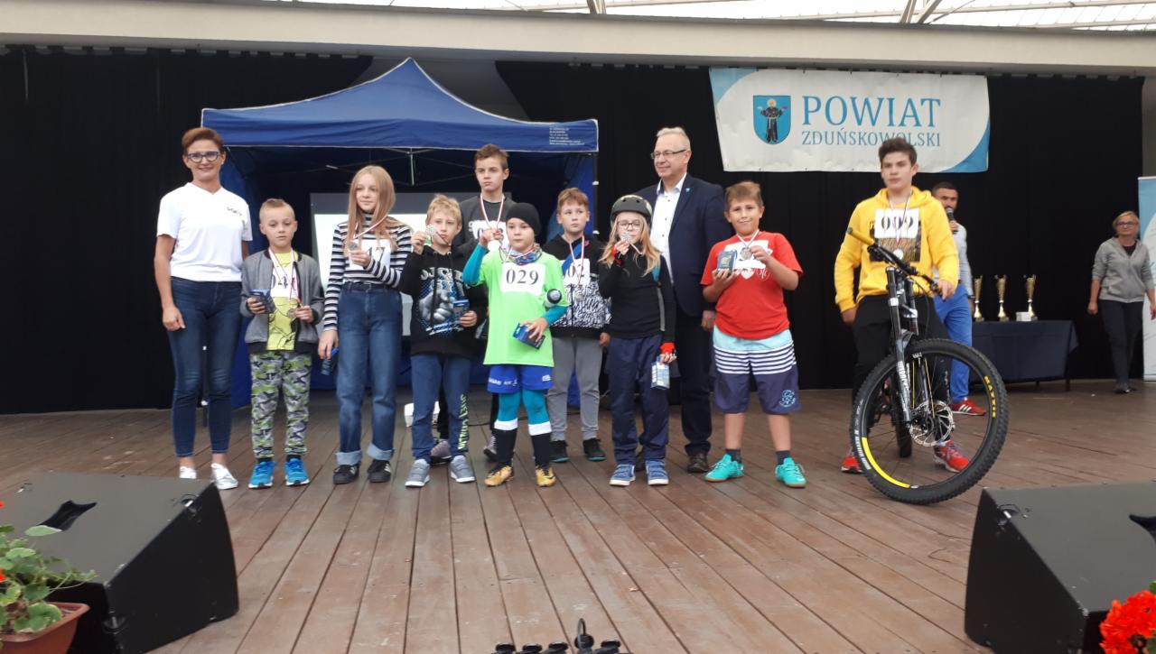 Grupa dzieci z medalami i nagrodami stoi na scenie podczas ceremonii wręczenia nagród sportowych, w tle widoczny baner Powiatu Zduńskowolskiego i rower.