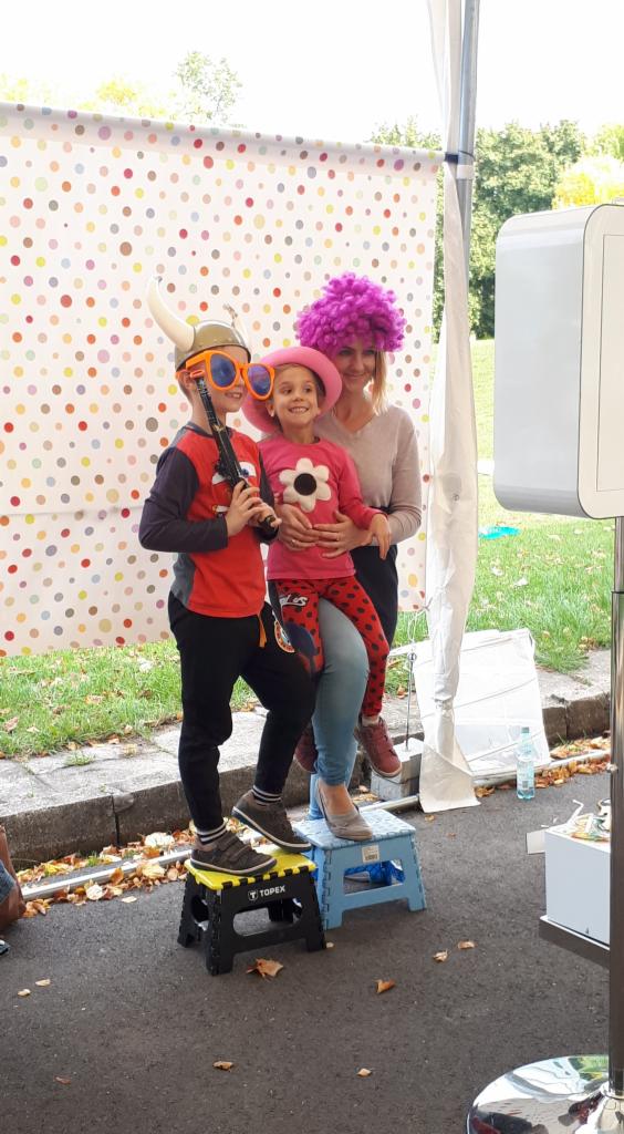 Rodzinne zdjęcie w fotobudce z kolorowym tłem w kropki, dzieci przebrane w zabawne rekwizyty: hełm wikinga, duże okulary i różową perukę, stoją na składanych stołkach.