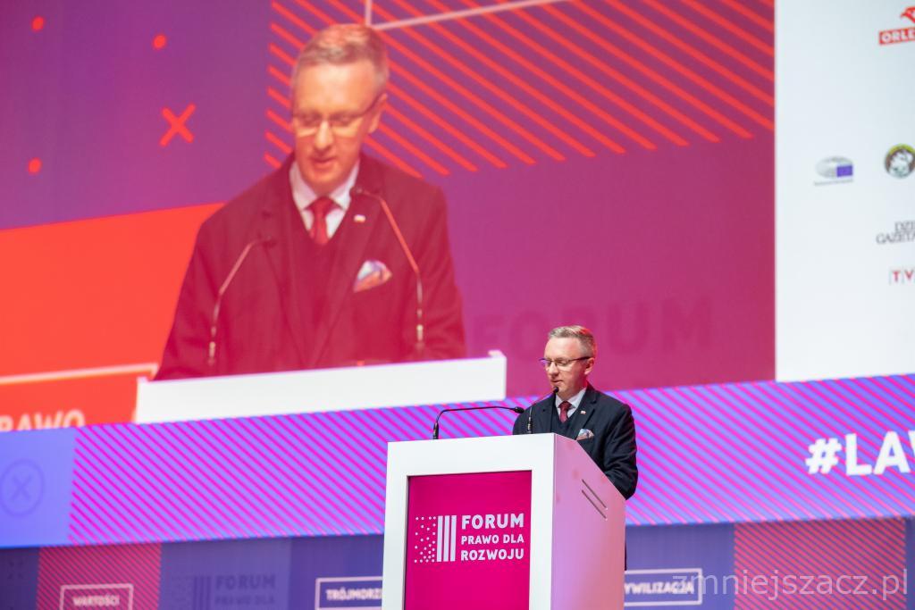 Prelegent w okularach z Łodzi przemawia na tle ekranu z logotypami podczas Forum Prawa dla Rozwoju, widoczny zbliżony kadr na ekranie.