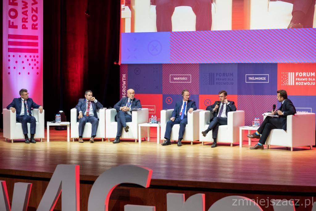 Panel dyskusyjny z udziałem pięciu mężczyzn w garniturach siedzących na białych fotelach na scenie z czerwonymi i niebieskimi elementami dekoracyjnymi w tle.