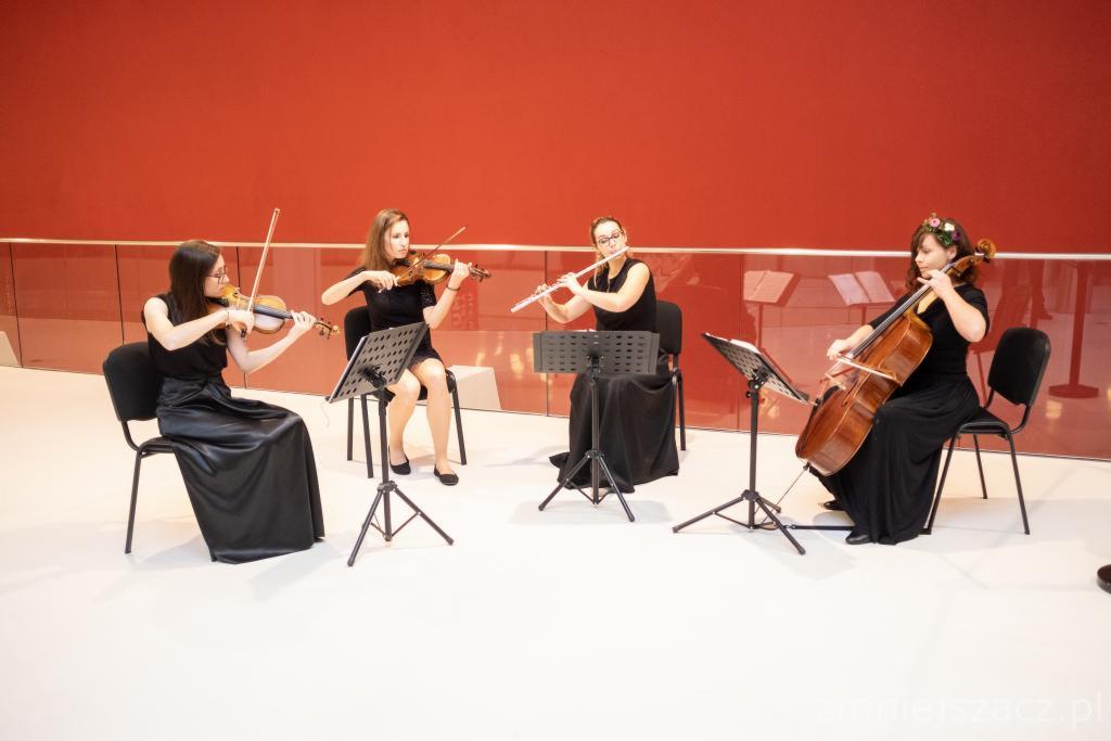 Kwartet smyczkowy w czarnych sukniach grający na skrzypcach, flecie i wiolonczeli na białej scenie z czerwonym tłem, nuty na pulpitach.