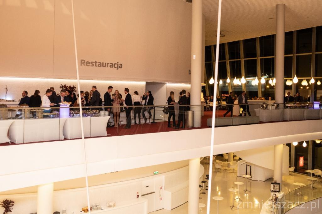 Wnętrze nowoczesnej restauracji z antresolą podczas przyjęcia, goście w strojach wieczorowych przy bufecie, widok z góry na minimalistyczne stoliki i krzesła na parterze.