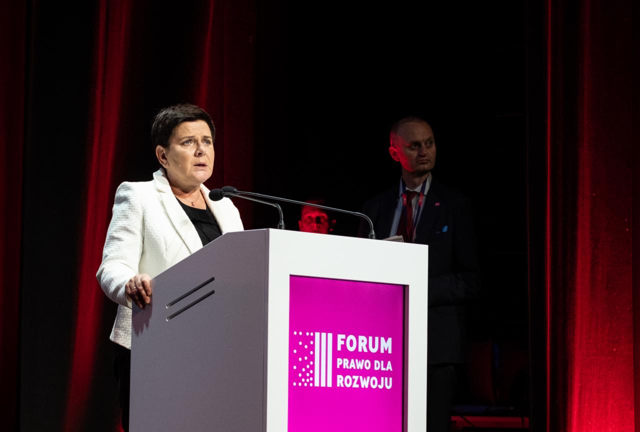 Kobieta w białym żakiecie przemawia z podium z logo Forum Prawo dla Rozwoju na tle czerwonych zasłon, w tle widoczny mężczyzna w garniturze.