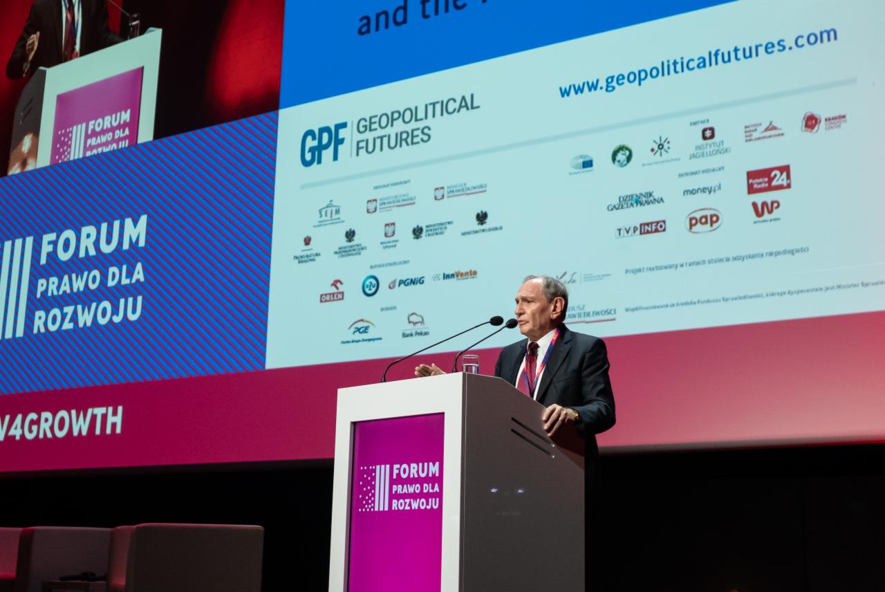 Prelegent w garniturze przemawia z podium podczas forum, na tle ekran z logo sponsorów, widać mikrofony i szklankę wody.