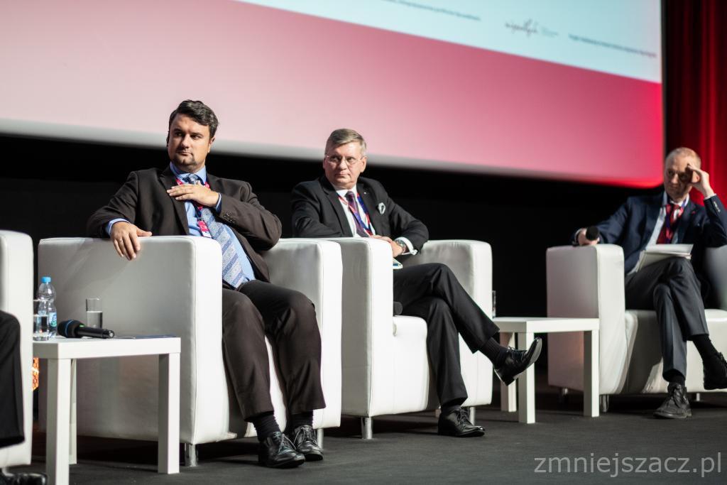 Trzej mężczyźni w garniturach siedzą na białych fotelach podczas panelu dyskusyjnego na tle ekranu projekcyjnego z wyświetlanym logotypem.
