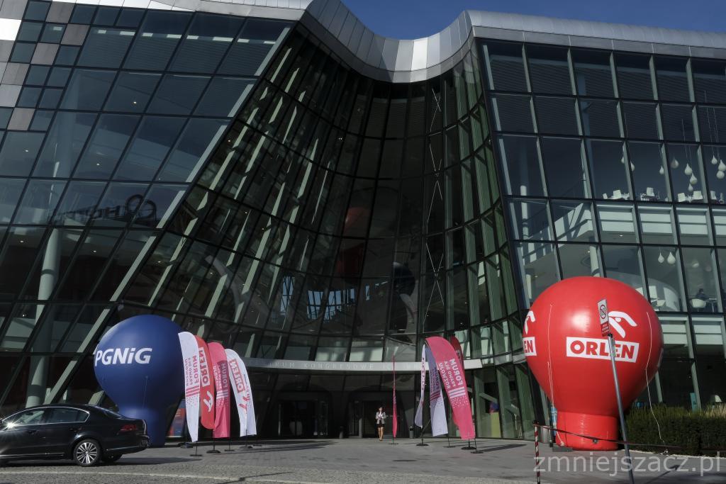 Nowoczesny budynek kongresowy z przeszkloną fasadą, udekorowany balonami reklamowymi PGNIG i Orlen oraz flagami promocyjnymi, przed wejściem zaparkowany czarny samochód.