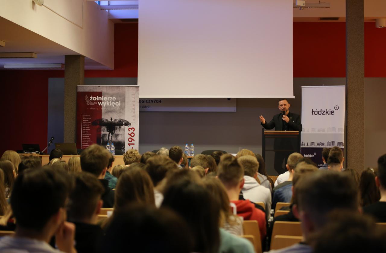 Prelegent w czarnym garniturze przemawia do zgromadzonego audytorium na tle ekranu projekcyjnego i banerów informacyjnych.