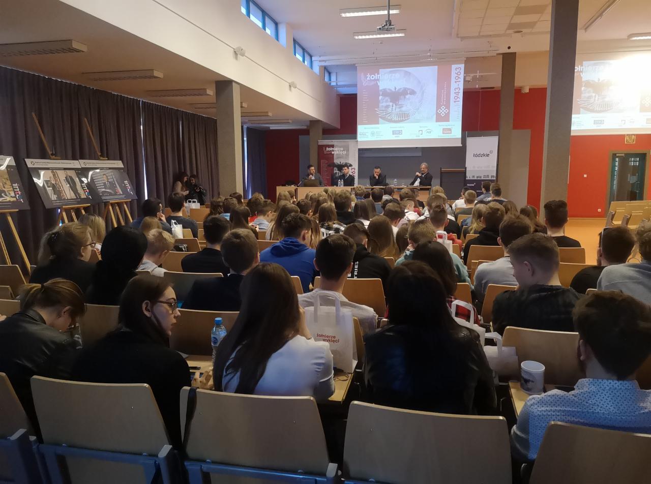 Widok z tyłu na salę konferencyjną wypełnioną uczestnikami słuchającymi prelegentów na scenie, z projekcją na ekranie w tle.