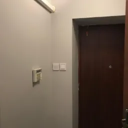 Wnętrze przedpokoju z zamontowanym panelem alarmowym, włącznikiem światła i brązowymi drzwiami wejściowymi.