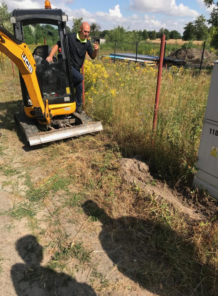 Operator w mini koparce JCB, z telefonem w dłoni, stoi obok skrzynki elektrycznej na tle łąki z kwitnącymi żółtymi kwiatami.