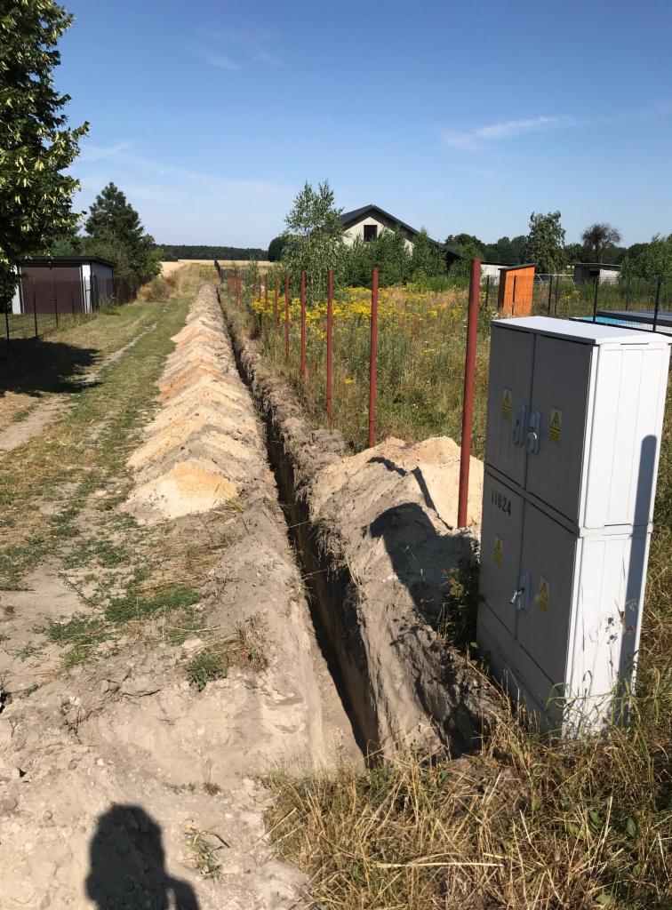 Wykopany rów w ziemi, prawdopodobnie pod kable elektryczne, obok skrzynka elektryczna z widocznymi ostrzeżeniami o napięciu, tło to łąka i zabudowania wiejskie.