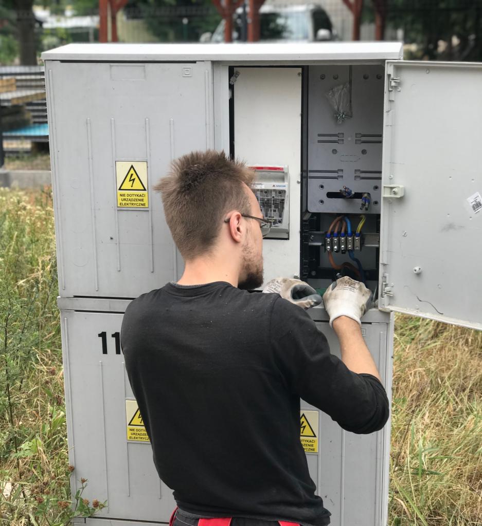 Elektryk w rękawicach roboczych, pochylony nad otwartą szafą elektryczną na zewnątrz, widoczne przewody i zabezpieczenia, tabliczka ostrzegawcza o napięciu.
