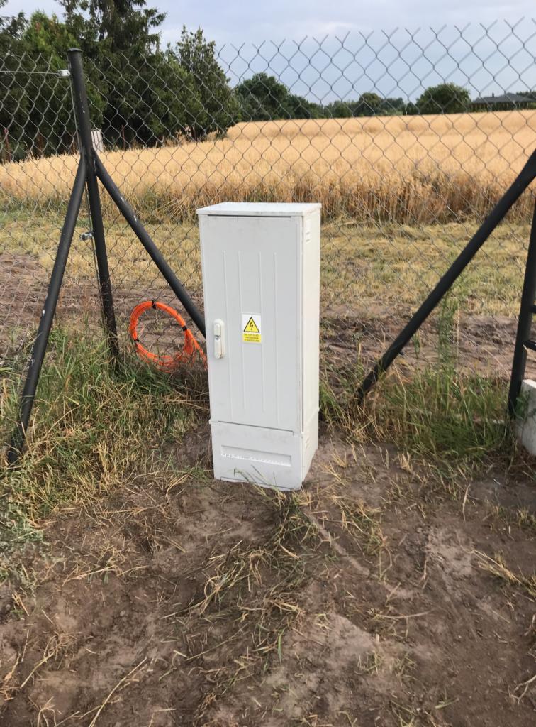 Zewnętrzna szafka elektryczna z ostrzeżeniem o napięciu, umieszczona na ziemi przy ogrodzeniu z siatki, z pomarańczowym kablem światłowodowym zwiniętym obok.
