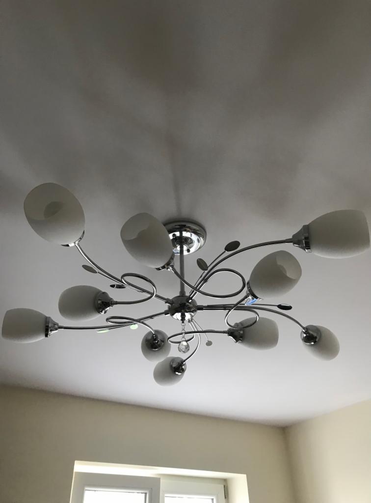 Chromowany żyrandol sufitowy z dziesięcioma mlecznymi, owalnymi kloszami i dekoracyjnymi elementami w kształcie liści, zawieszony na białym suficie z widocznym oknem w tle.