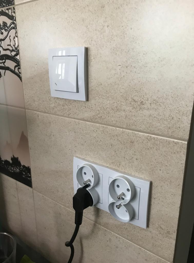 Biały włącznik światła i podwójne gniazdko elektryczne z czarną wtyczką, zamontowane na ścianie wyłożonej płytkami w odcieniach beżu.