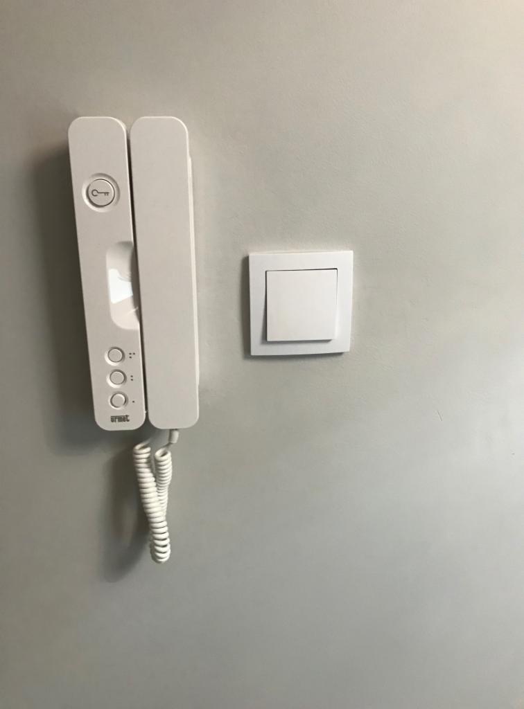 Biały domofon marki Urmet z przyciskiem klucza i trzema przyciskami dzwonków, zamontowany obok białego włącznika światła na szarej ścianie.