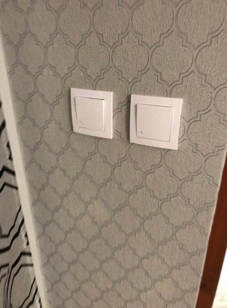 Dwa białe, kwadratowe włączniki światła na ścianie pokrytej tapetą w szare wzory koniczyny marokańskiej, widoczne z bliska.