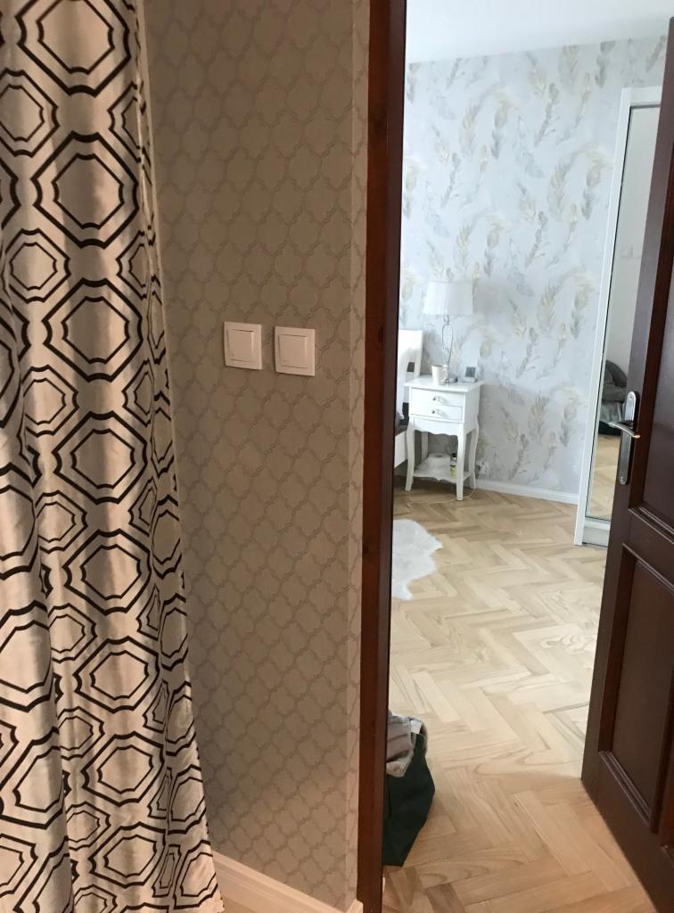 Dwa białe gniazdka elektryczne zamontowane na ścianie z wzorzystą tapetą, widoczne obok zasłony i framugi drzwi prowadzących do sypialni z parkietem w jodełkę i białą szafką nocną.