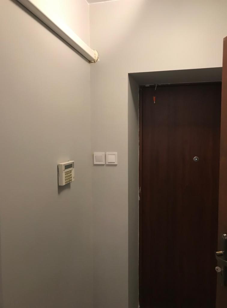 Wnętrze przedpokoju z zamontowanym panelem alarmowym, włącznikiem światła i brązowymi drzwiami wejściowymi.