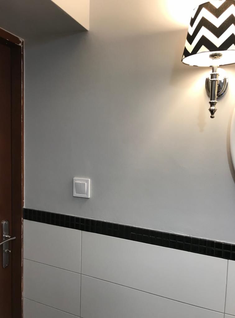 Ściana w łazience z włącznikiem światła, lampą ścienną z abażurem w zygzaki i fragmentem drzwi z klamką.