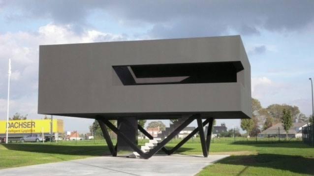 Nowoczesny budynek o kubistycznej bryle, uniesiony na skośnych podporach, z minimalistycznym oknem i schodami, na tle pochmurnego nieba i zielonego krajobrazu.