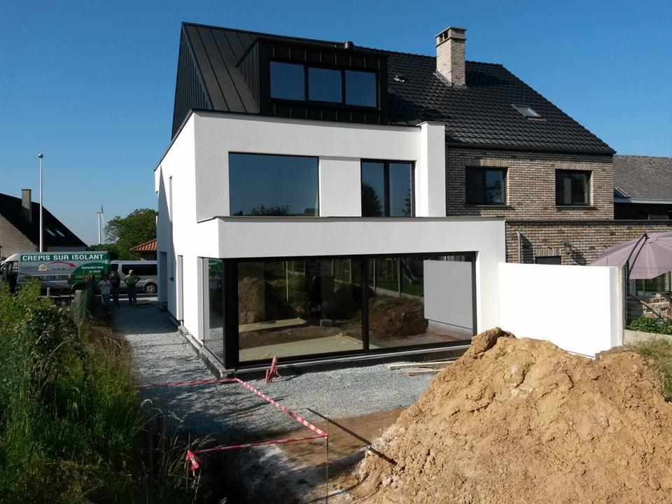 Nowoczesny, dwukondygnacyjny dom z białym tynkiem i czarnym dachem, w trakcie prac wykończeniowych elewacji, widoczny stos ziemi i ekipa przy samochodzie z napisem 'Crepis sur Isolant'.