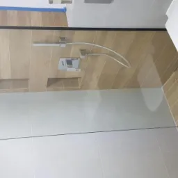 Nowoczesna kabina prysznicowa ze szklaną ścianką i deszczownicą, wykończona płytkami imitującymi drewno i szarymi płytkami, z widocznym wiszącym sedesem.