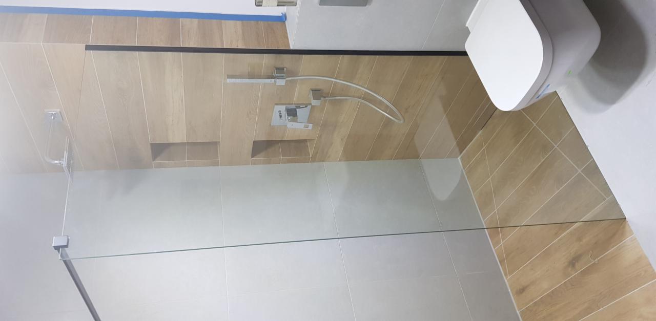 Nowoczesna kabina prysznicowa ze szklaną ścianką i deszczownicą, wykończona płytkami imitującymi drewno i szarymi płytkami, z widocznym wiszącym sedesem.