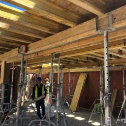 Wnętrze w trakcie budowy, widoczne szalunki stropowe wsparte na stalowych podporach, mur z czerwonej cegły oraz dwóch pracowników w odzieży roboczej i kaskach.