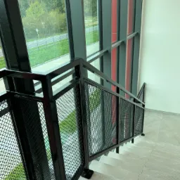 Nowoczesne schody z czarną metalową balustradą z siatki, widok z góry, duże okno z widokiem na zieleń w tle.