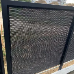 Częściowy widok czarnego, perforowanego panelu ogrodzeniowego z metalową ramą, na tle placu budowy z budynkami mieszkalnymi i żurawiem.